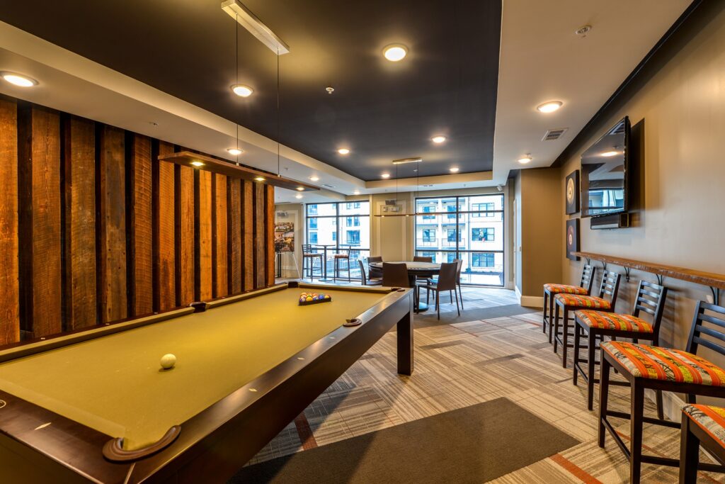 Community center with pool table, bar stools, wooden accent wall, and tables and chairs. Mounted tv on the wall.