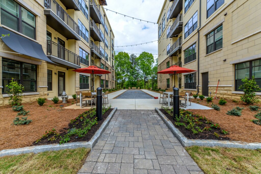 Walking area between 2 residential buildings with string lights and 2 tables with umbrellas and chairs. Bocce ball court in the middle of walk way.