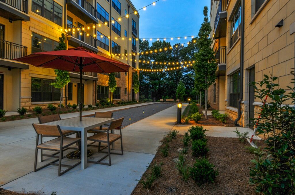 Out door seating area with string lights and umbrellas. Seated between 2 residential buildings. Bocce ball court in the back.
