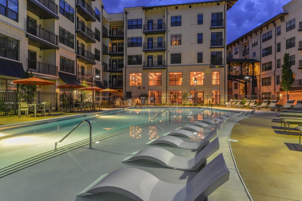 Night view of in pool seating with pool view. Lounge chairs on the right of the outside of the pool and tables with umbrellas and chairs on the left.