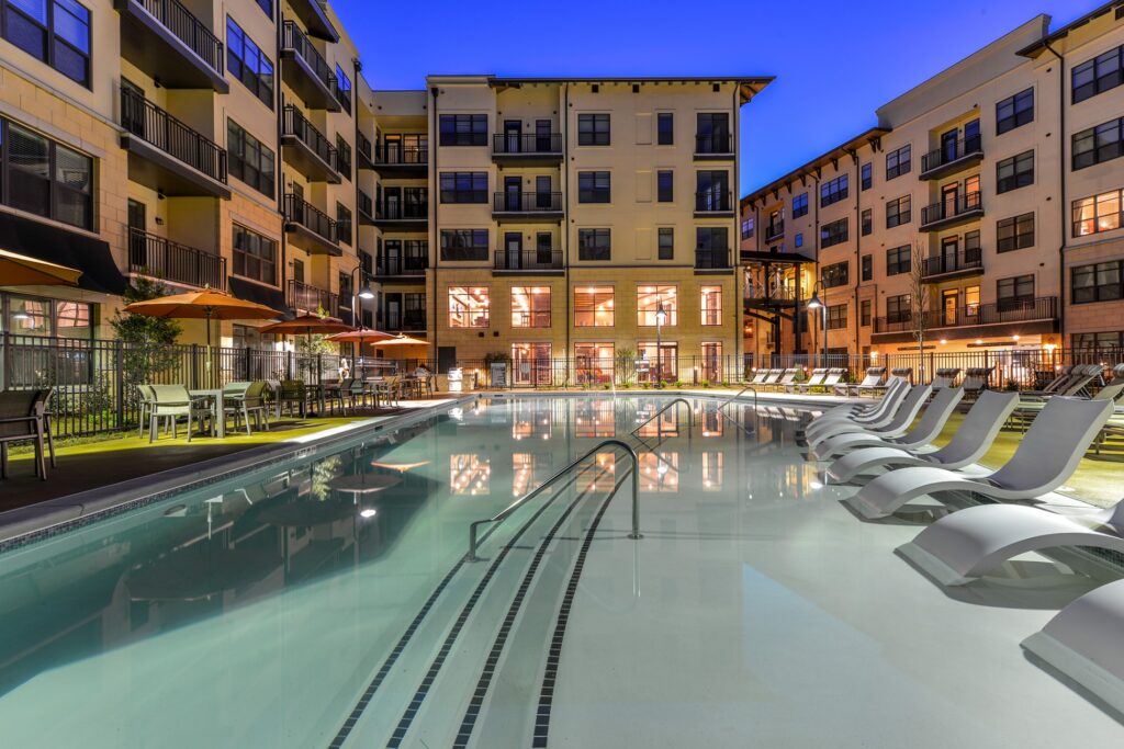 Night view of in pool seating with pool view. Lounge chairs on the right of the outside of the pool and tables with umbrellas and chairs on the left.