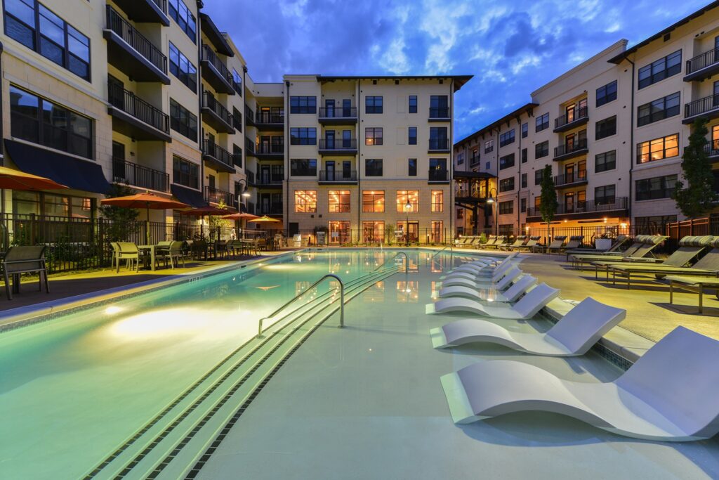Night view of in pool seating with pool view. Lounge chairs on the right of the outside of the pool and tables with umbrellas and chairs on the left.