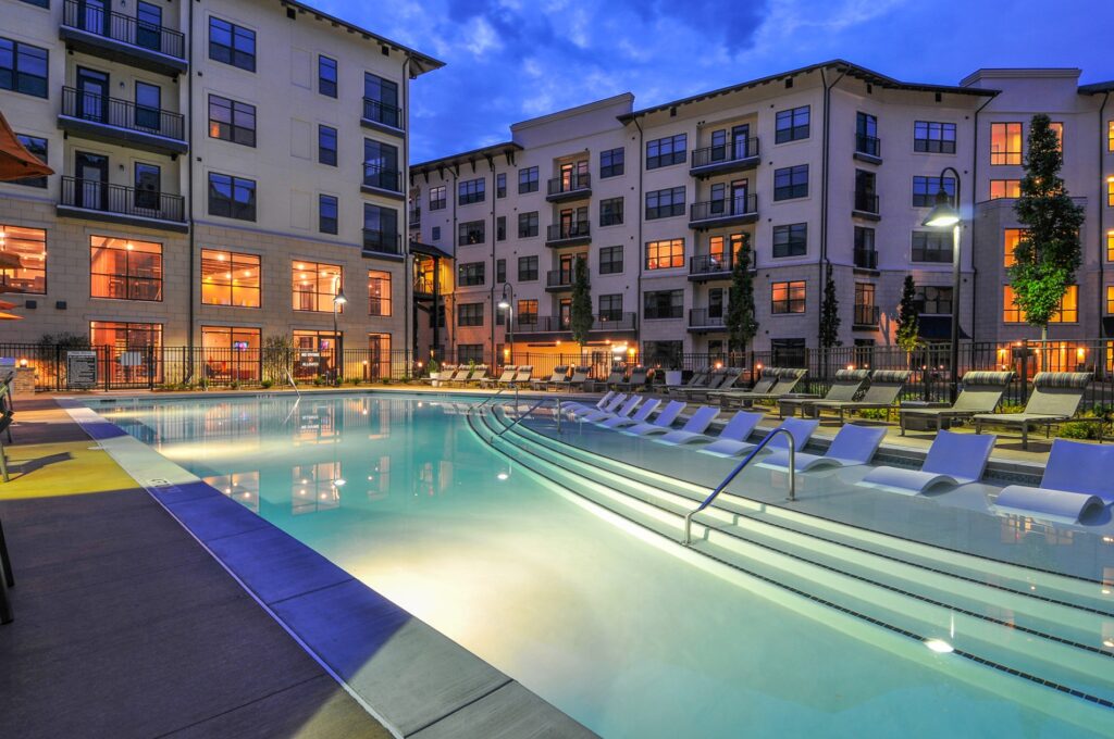 Night view of in pool seating with pool view. Lounge chairs on the right of the outside of the pool and tables with residential buildings in the back.