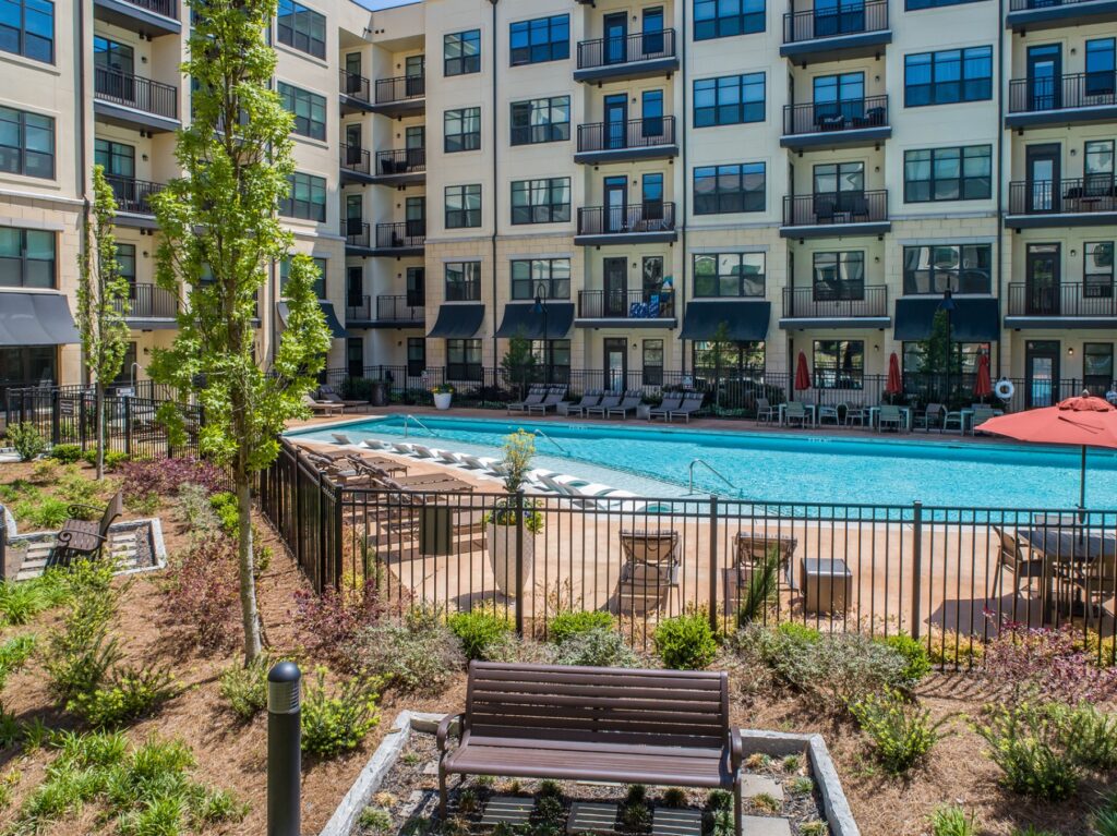 Gated pool area with lounge chairs, in pool seating, umbrella tables and chairs. Benches around the pool. With residential building balconys in the back